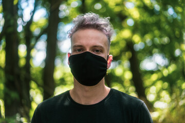 portrait of young man with white hair wearing a black face mask in forest in summer during corona