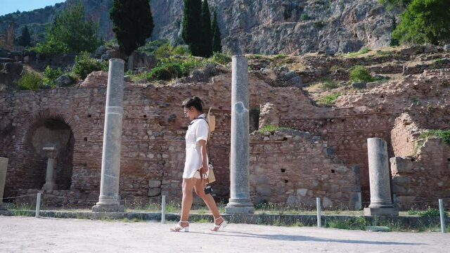 Young woman visiting ruins of temple of Apollo in Delphi, archaeological site by mount Parnassus, Greece, Europe. Large hat, fashion white dress.
