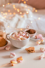 Ceramic cup of hot chocolate or cocoa with marshmallow on white table. Warming Christmas winter drink. Decoration garlands of lights.