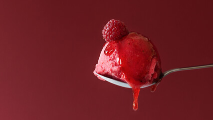 A scoop of raspberry ice cream with melting raspberry topping on top over red background.