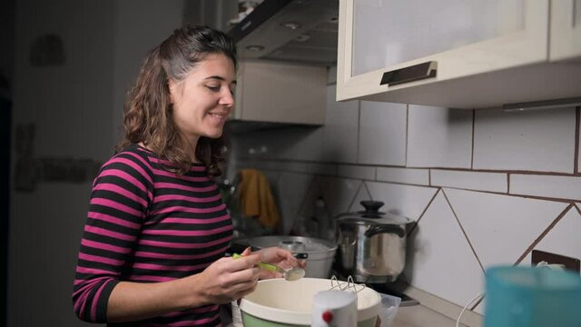 Medium-shot smiling caucasian woman cooking in her kitchen. Making a cake, putting ingredients in a bowl to prepare the dough. Cooking hobby, life at home concept.