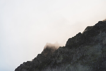 Dramatic bleak fog among giant rocky mountains. Ghostly atmospheric view to big cliff in cloudy sky. Low clouds and beautiful rockies. Minimalist scenery in mysterious place at early foggy morning.
