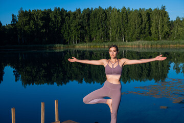 Fototapeta na wymiar Young woman by the lake practicing yoga moves on wooden platform. Pretty young woman exercising in nature, healthy lifestyle young people positive vibes.