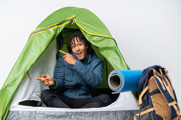 Young african american man inside a camping green tent surprised and pointing side