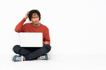 Young african american man sitting on the floor and working with his laptop having doubts while scratching head