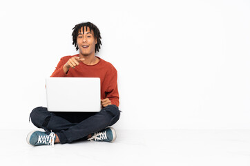 Young african american man sitting on the floor and working with his laptop surprised and pointing front