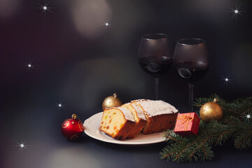 Christmas cake with candied fruits and raison, two glasses of red wine, fir branches and christmas decorations on dark background.