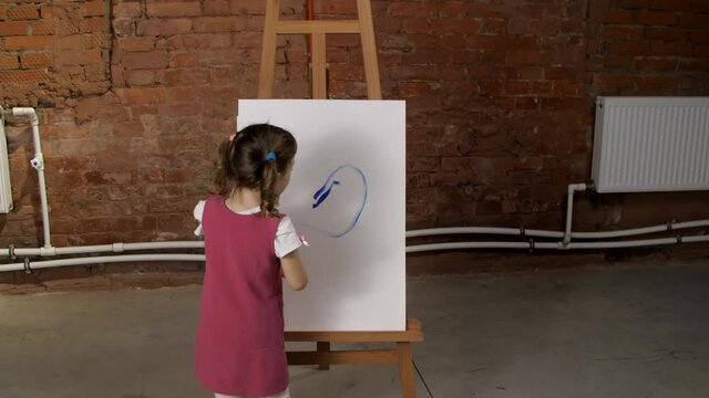 Pretty little three year old girl draws with blue paint on canvas on an easel, finishing up her work, adding little details to the painting. Children's creativity. Close-up shot of painting process. 