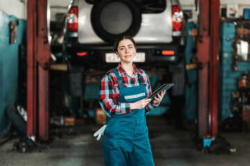A young smiling mechanic female poses in a uniform with a tablet in her hands. In the background, a car on a lift. Technical inspection and diagnostics of the car using modern technologies