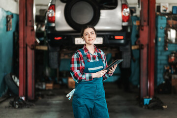 A young smiling mechanic female poses in a uniform with a tablet in her hands. In the background, a...