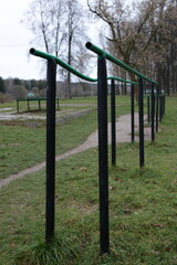 Fototapeta premium Empty old bent metal gymnastic bars on an abandoned sports field in the Park on a cloudy autumn day on green grass and bare trees background