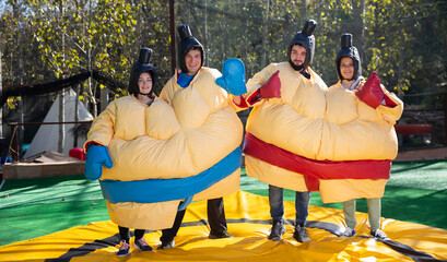 Portrait of happy adult friends having fun posing in inflatable sumo suits at outdoor amusement park - obrazy, fototapety, plakaty