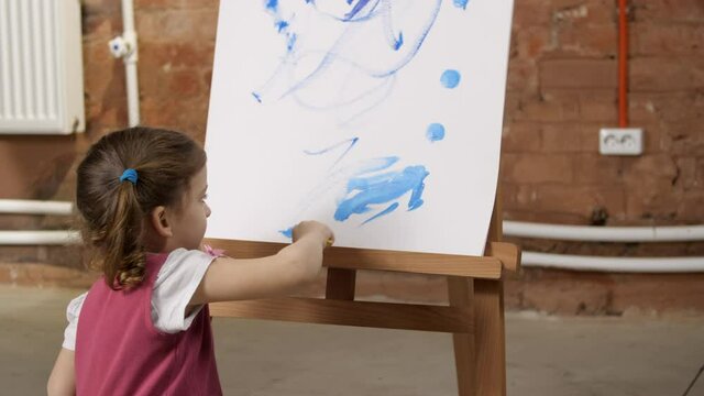 Pretty little three year old girl draws with blue paint on canvas on an easel, finishing up her work, adding little details to the painting. Children's creativity. Close-up shot of painting process.