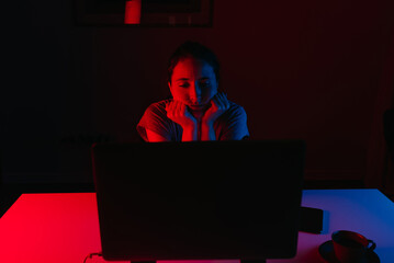 A woman is working remotely on a laptop at home. A girl is resting her head on her hands which is leaning on the table. A female employee is bored in front of the computer under blue and red light.