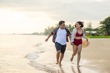 Smiling young Asian couple tourist holding hands together and enjoy running on beach at summer sunset. Happy man and woman relax and having fun with outdoor lifestyle in summer holiday vacation.