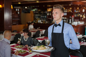 Portrait of smiling waiter with serving tray meeting restaurant guests ..