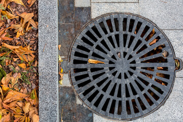 manhole cover round black metal lattice on a city park pedestrian sidewalk paved with granite gray tiles with a stone border close-up top view, nobody.