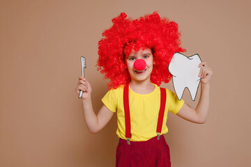 Funny clown girl child in a red wig and with a nose holding a mock-up of a tooth and brush isolated against the background of a Set Sail Champagne flower. Kid teeth hygiene, teeth cleaning concept.
