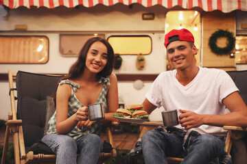 Happy couple with burgers and cups resting near trailer. Camping season