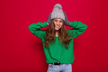 Lovable european girl expressing happiness. Refined long-haired young woman in green sweater and grey knitted cap laughing on red background in new year.