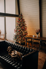 Large Christmas tree with warm lights in the living room with a large panoramic window. Cozy living room in a loft style with a large terrace. Vertical photo