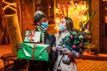 Close up portrait of joyful lovely mixed-races couple standing in evening in city with christmas presents in medical masks. Man and woman on snowy street looking at camera and smiling. Xmas concept