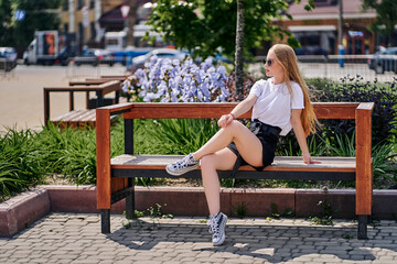 A teenage girl sits on a bench in a city park. Turning his head looks into the distance.