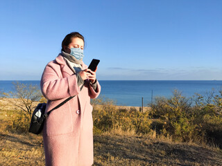 A girl in a medical mask looks at the phone against the background of the autumn sea