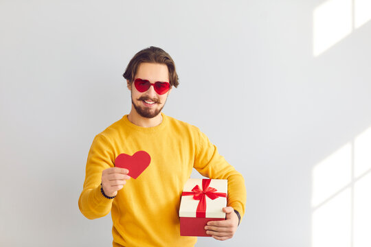 Smiling young man in heart shaped sunglasses with present box showing red heart during online dating