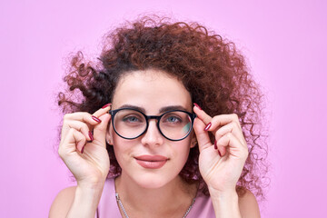 Armenian girl with curly hair holds hands on the rim of glasses and squints, trying to see...
