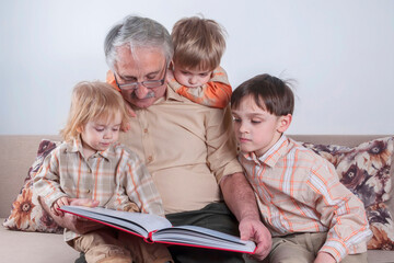 Positive elderly man reading a book for his grandchildren. Three children listen with interest