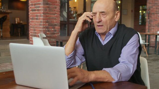 Old italian businessman talks on phone and works on notebook in cafe