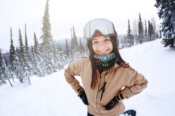 Beautiful girl snowboarder smiling, looking at the camera, around the snowy forest of the ski resort.