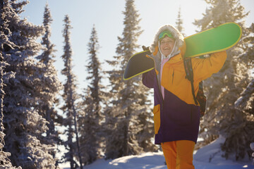 A snowboarder with a snowboard on her shoulders in the winter forest, the sun is shining from behind.