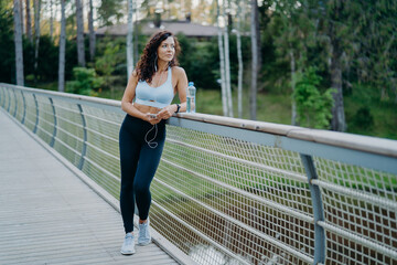 Fototapeta premium Serious thoughtful woman with spoty body, dressed in cropped top and leggings, poses at bridge looks pensively away, listens music in earphones. People, healthy lifestyle and fitness concept