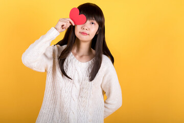 Portrait of lovely asian woman in white sweater showing heart romantic symbol and looking at camera with smile, holding valentine's day greeting card. studio shot isolated on yellow background.