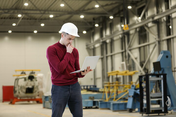 Portrait of a male factory manager in a white hard hat and red sweater holding laptop and mobile phone. Controlling the work process in the helicopter manufacturer.	