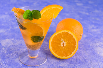 A glass of refreshing orange cocktail with mint and ice on a blue bright background.	