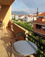 Balcony with gray table and bronze chairs in the house. In the background, a rainbow over the mountain and white houses with red tiles. Budva  Montenegro