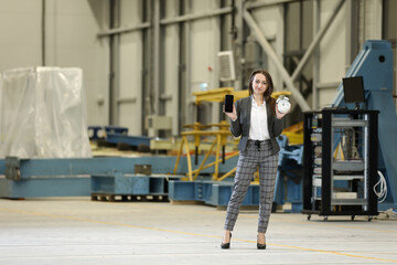 Portrait of a female factory manager in a business suit holding mobile phone and retro alarm clock , controlling the work process in the helicopter manufacturer. Timeline. Deadline. 