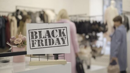Close-up of Black Friday announcement in clothing store with blurred women choosing clothes at the background. Wealthy Caucasian shopaholics shopping in pricey mall. Shopaholism concept.