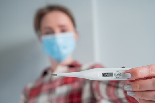 Portrait of woman in medical face mask showing digital medical thermometer with high temperature - selective focus. Healthcare, measurement, disease, covid 19, infection, coronavirus concept