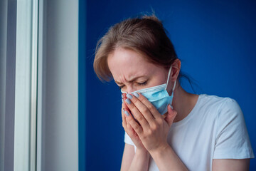 Woman wearing face mask, feeling sick, suffering from sneezing, looking out of window in room with blue wall. Self isolation, prevention, quarantine, COVID-19, coronavirus, safety concept