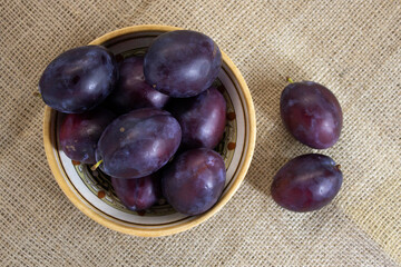 Big purple plums in a ceramic dish with ethnic ornament on a rough linen background, autumn harvest, top view, close up