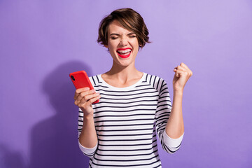 Photo of amazed crazy girl hold smartphone raise fist closed eyes wear striped shirt isolated purple color background