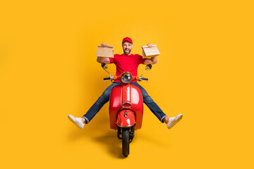 Photo of careless crazy guy drive bike hold two packages wear red t-shirt cap jeans isolated yellow color background © deagreez