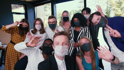 Group of happy diverse business colleagues posing, waving for selfie photo in masks against COVID-19 at modern office.