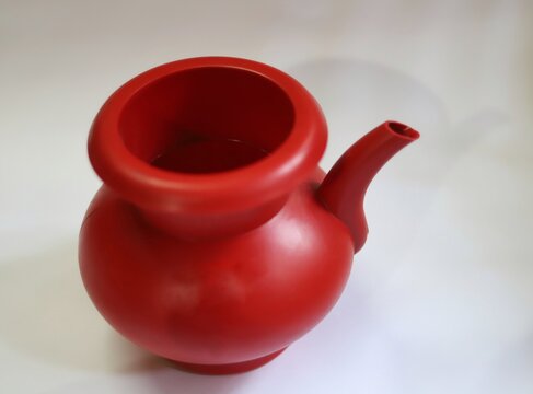A close up view of a red plastic lota or "bodna" or watering pot placed on a white surface . It is a type of spouted vessel made of brass, copper or plastic used in Indian subcontinent.