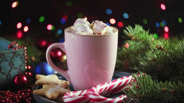 Male hand puts a cup of fragrant, hot cocoa with marshmallows on a table with branches of a Christmas tree and glowing garland lights. Preparing and serving a hot Christmas drink.