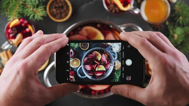 Male hand takes pictures with a smartphone composition with hot wine drink with spices and fruits, on a dark background with Christmas tree branches. Fragrant, hot punch or mulled wine for Christmas.